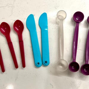 May include: An assortment of colorful plastic kitchen utensils, including three red spoons, two blue spreaders, a clear measuring spoon, and two purple measuring spoons. The utensils are arranged on a white surface.