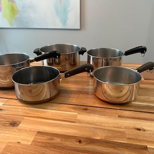 Könnte beinhalten: Ein Set aus fünf Edelstahl-Kochtöpfen mit kupferfarbenen Böden und schwarzen Griffen. Die Kollektion umfasst Töpfe in verschiedenen Größen, die jeweils über eine polierte Metalloberfläche, einen kreisförmigen Rand und einen stabilen Griff mit einer kleinen Metallöse zum Aufhängen verfügen.