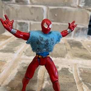 以下が含まれることがあります： 雪の結晶模様の青いシャツを着た、赤と青のスパイダーマンのアクションフィギュア。フィギュアは腕を伸ばし、足を広げてポーズをとっている。