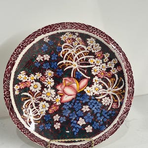 Könnte beinhalten: Ein antiker Keramikteller mit Blumenmuster. Der Teller hat einen burgunderfarbenen Rand mit einem weißen Blumenmuster auf dunkelgrünem Hintergrund. In der Mitte des Tellers befindet sich eine rosa Blume, umgeben von weißen und blauen Blumen.