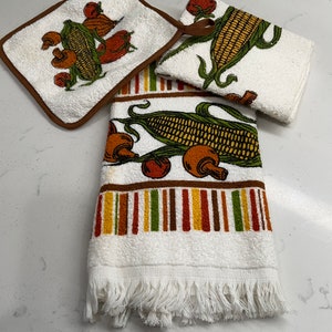 Op de afbeelding: Set van drie keukendoeken met een witte achtergrond en een bruin, geel, groen en rood maïs- en paddenstoelontwerp. De handdoeken hebben een franje rand.