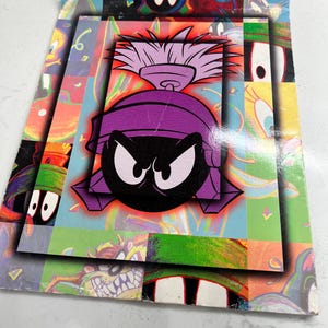 May include: Stacked, colorful Looney Tunes cards featuring Marvin the Martian. The top card has a large image of Marvin's face, with a purple helmet and black eyes. The background is a mix of bright colors and cartoon characters.