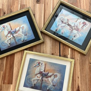 May include: Three framed pictures of a white carousel horse with gold frames. The pictures are on a wooden surface.
