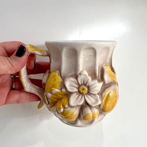 Könnte beinhalten: Eine cremefarbene Keramiktasse mit floralem Design. Die Tasse hat eine weiße Blume mit gelber Mitte und gelben Blättern. Der Griff ist ebenfalls cremefarben mit einem astähnlichen Design.