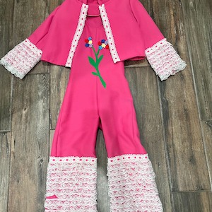 May include: A pink jumpsuit with a matching pink cape. The jumpsuit has white lace trim on the legs and sleeves. The cape has white lace trim around the edges. The jumpsuit has a flower design on the front.