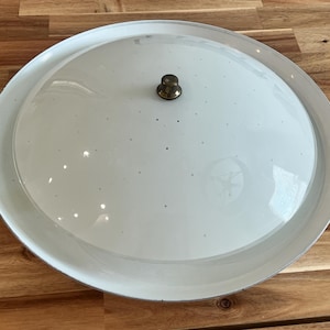 May include: A round, white ceiling light fixture with a brass-colored center knob. The fixture has a slightly domed shape and a white rim. The light is set against a wooden surface.