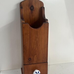 Puede incluir: Un portafósforos de madera vintage para colgar en la pared, con un cajón. El soporte es de madera teñida oscura y presenta una parte superior decorativa con un agujero. El cajón tiene un pomo de cerámica blanco y azul con motivos florales. Una pieza encantadora para una cocina rústica.