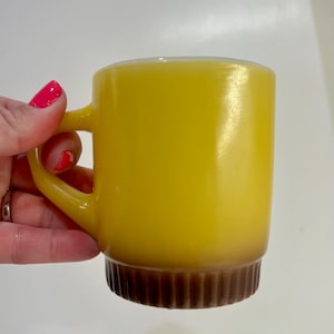 Op de afbeelding: Een gele glazen mok met een glad bovenlichaam en een donkerbruine, verticaal geribbelde basis. De mok heeft een C-vormig handvat aan de zijkant, met een doorschijnende afwerking waardoor licht door het gele glasmateriaal heen kan schijnen.