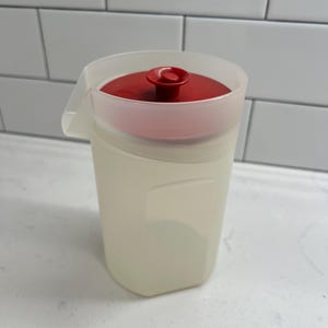 Op de afbeelding: Een doorschijnende plastic kan met een rode deksel en handvat. De kan heeft een rechthoekige vorm en een tuit om uit te schenken. De deksel heeft een kleine, afgeronde handgreep. De kan is ontworpen voor het vasthouden en serveren van vloeistoffen.