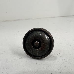 以下が含まれることがあります： 円形のデザインの、暗いアンティークスタイルのキャビネットノブ。ノブは、わずかにテクスチャ加工された表面と、中央に隆起した円形のディテールを備えた暗い金属製です。 家具やキャビネット用です。