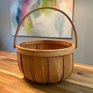 May include: A round, light brown wooden basket constructed from vertical slats with a reinforced rim and a single, arched handle attached to the sides. The basket features a natural wood grain finish and is held together by small metal fasteners.