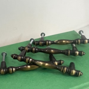 May include: A set of four antique-style brass drawer pulls with a dark, tarnished finish and polished gold-tone highlights. Each handle features a central contoured grip, decorative ribbed detailing, and two threaded metal mounting screws.