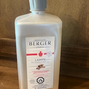 Könnte beinhalten: Eine durchsichtige Kunststoffflasche mit Maison Berger Paris Lampen-Nachfüllflüssigkeit im Duft Christmas Cookies. Das Etikett zeigt den Markennamen, ein rotes Symbol, eine Schneeflocke und den Text 1L-33.8 US fl.oz. mit einem Gefahrenhinweis.