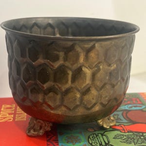 Pode incluir: Um vaso redondo de latão metálico com um padrão de favo de mel texturizado na superfície exterior. O recipiente é suportado por três pés pequenos e ornamentados, apresentando um acabamento de pátina escura e envelhecida.
