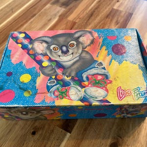 May include: A rectangular box with a colourful design featuring a cartoon koala holding a polka-dotted item. The box has a blue background with pink and yellow accents, and the words "Lisa Frank" are visible.