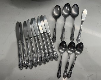 Puede incluir: Un conjunto de cubiertos antiguos, que incluye cuchillos, cucharas y un cuchillo para mantequilla. La cubertería está hecha de un metal brillante de color plateado y presenta detalles florales ornamentados en los mangos. El conjunto está dispuesto sobre una superficie clara.