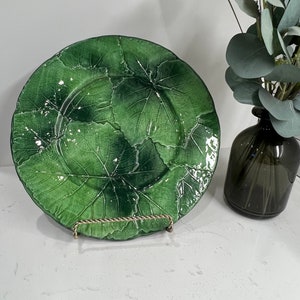 Pu&ograve; includere: Un piatto da portata in ceramica verde con un motivo a foglie. Il piatto &egrave; su una superficie bianca ed &egrave; sostenuto da un supporto in metallo.