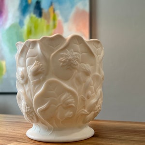 Puede incluir: Jardinera de cerámica blanquecina con un diseño detallado de flores y hojas. La jardinera tiene una parte superior ancha y abierta y una base de pedestal. El patrón en relieve incluye flores y hojas, creando una superficie texturizada.