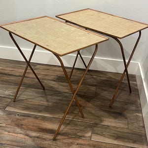 May include: A pair of rectangular folding snack tables featuring light tan woven cane tops framed in wood. Each table is supported by thin, dark brown metal legs that cross in an X-shape, providing a stable base for these portable furniture pieces.