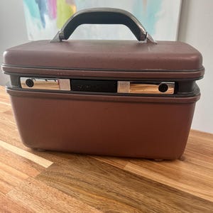 Puede incluir: Una maleta de viaje vintage marrón con asa negra y detalles plateados. La maleta tiene la palabra "Samsonite" impresa en la parte delantera.