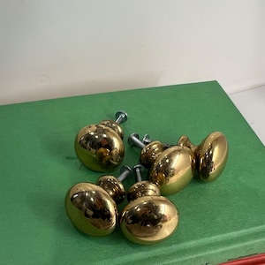 May include: A collection of shiny, gold-colored cabinet knobs with silver screws. The knobs are oval-shaped and reflect light, resting on a green surface. Hardware for furniture.