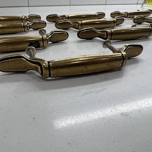 May include: A collection of brass-toned metal drawer pulls featuring a central horizontal bar and two side mounting points. Each pull includes two threaded metal screws extending from the back. A yellow measuring tape is visible in the corner.