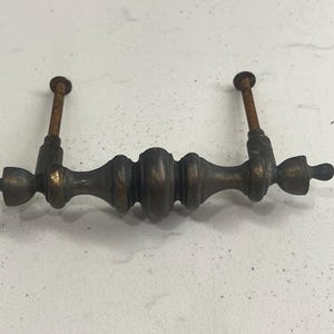 Puede incluir: Un tirador de armario de estilo vintage de bronce oscuro con un diseño redondeado y ornamentado. El tirador tiene dos agujeros para tornillos en cada extremo.