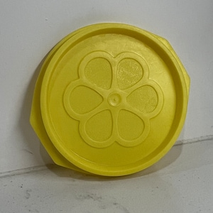 Puede incluir: Tapa de plástico amarilla con un diseño de flor. La tapa es redonda y tiene un borde ligeramente elevado.