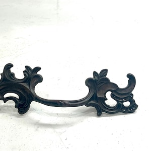 May include: Ornate bronze-colored drawer pull with a decorative, scrolling design. The handle features a central bar with elaborate floral and foliate details on each end. A vintage-style hardware element.