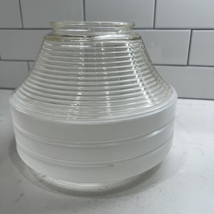 May include: Clear glass and white light fixture with a ribbed, cone-shaped top and a rounded, white base. The fixture is designed with horizontal ridges and a smooth, rounded bottom. The glass top is transparent, and the base is opaque white.