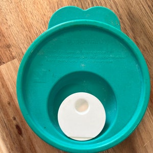 Peut inclure: Un couvercle Tupperware bleu turquoise avec une aération blanche au centre. Le couvercle porte l'inscription "Tupperware" et le texte "Green for use in microwave, Ouvrir avant usage au micro-ondes, Abrir para uso en microondas".