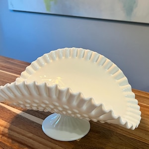 May include: A white milk glass bowl with a ruffled edge and a pedestal base. The bowl is decorated with small raised dots.