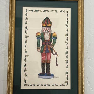 May include: A framed watercolor painting of a nutcracker soldier wearing a crown, green jacket, and red trousers, holding a sword. The artwork is bordered by a decorative holly leaf and berry pattern on a cream background, encased in a gold frame.
