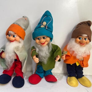 Könnte beinhalten: Drei Zwergenfiguren mit weißen Bärten und spitzen Mützen. Die erste trägt eine graue und orangefarbene Mütze mit grauem Umhang und roter Hose. Die zweite trägt eine blaue Mütze mit grüner Jacke und blauer Hose. Die dritte trägt eine braune Mütze mit orangefarbener Jacke und dunkelblauer Hose.