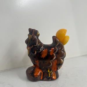 May include: A ceramic hen figurine with a brown, orange, and yellow glaze. The hen has a detailed, textured body and a bright yellow comb and wattle. The figurine is a decorative item.
