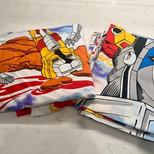 Puede incluir: Dos sábanas blancas dobladas con ilustraciones coloridas de personajes de Voltron. Una sábana muestra un robot león amarillo, la otra una cara de robot con acentos azules, rojos y grises. Las sábanas tienen una estética vintage.