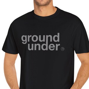 May include: Black t-shirt with the text "ground under" in gray, featuring a round trademark symbol. The shirt is a crew neck style and is being worn by a person in blue jeans.