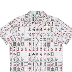 May include: A white short-sleeved button-down shirt with a pattern of mahjong tiles. The tiles are in black, red, and green, and they feature Chinese characters and numbers.