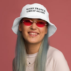 Puede incluir: Sombrero de pescador blanco con el texto "MAKE TRANCE GREAT AGAIN" en negro. La persona lleva gafas de sol tintadas de rojo y una camiseta de manga larga de color claro. El fondo es de color rosa liso.