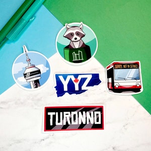 May include: Five colourful stickers featuring Toronto landmarks and symbols. The stickers include a raccoon wearing a green jacket, a red bus with the text "Sorry, Not in Service", a blue and white sticker with the text "YYZ", a white sticker with a tall tower, and a red and black sticker with the text "TORONTO".