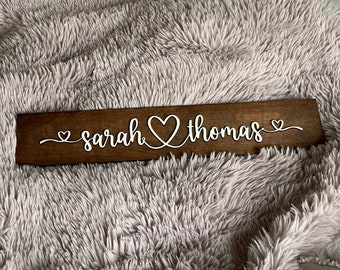 May include: A rectangular dark brown wooden sign featuring the white script text "sarah" and "thomas" connected by a central heart shape. Smaller heart icons are positioned at the beginning and end of the names, creating a continuous design.