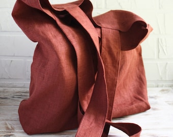 May include: A rust-colored linen tote bag with long straps. The bag is made of a textured fabric and has a simple, minimalist design. The straps are long enough to be worn over the shoulder. The bag is sitting on a light-colored wooden surface.
