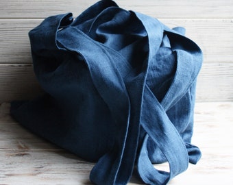 May include: A blue linen tote bag with long straps folded over the top. The bag is sitting on a white wooden surface.