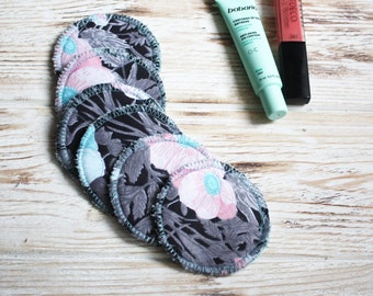 May include: Six black and white reusable makeup remover pads with floral print and teal stitching. The pads are stacked on a white wooden surface. A tube of eye cream and a tube of lip gloss are also visible.