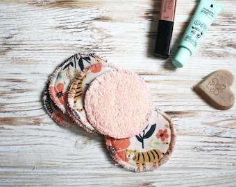 May include: A set of reusable makeup remover pads with a pink terrycloth side and a patterned cotton side featuring tigers and flowers. The pads are arranged in a stack on a white wooden surface. There are also two tubes of makeup remover and a heart-shaped bar of soap in the image.