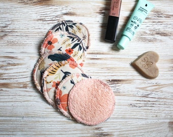 May include: A set of six reusable makeup remover pads. Five pads are made of cotton fabric with a floral print and one pad is made of a soft pink terry cloth. The pads are stacked on a white wooden surface. There is also a tube of face cream, a tube of lip gloss, and a heart-shaped bar of soap in the image.