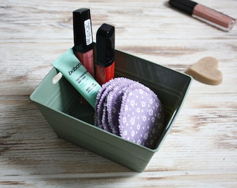 May include: A green metal basket with two black lip glosses, a small tube of eye cream with the text "babaria contorno de ojos anti edad", and a stack of purple cotton rounds with a white floral pattern.
