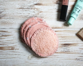 May include: A stack of reusable, round, peach-colored makeup remover pads. Some pads have a soft, textured surface, while others have a floral pattern. The pads are arranged on a light-colored, wooden surface.