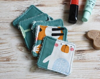 May include: A stack of reusable, square face cloths with teal backing and various printed designs. One cloth features a white bear with blue accents, another has an orange and black tiger pattern. Other cloths have green and white patterns. The cloths are on a light wood surface.