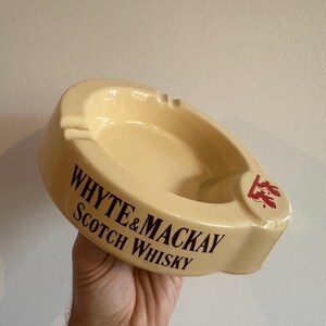 Op de afbeelding: Crèmekleurige keramische asbak met de tekst "WHYTE & MACKAY SCOTCH WHISKY" in donkerbruine letters. De asbak heeft een afgeronde vorm met een klein rustgedeelte voor sigaretten en een rood logo.